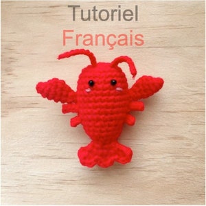 Peut inclure: Un homard en crochet rouge avec des yeux noirs et des joues roses. Le homard est sur une surface en bois. Le texte "Tutoriel Français" est au-dessus du homard.