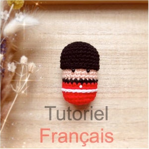 Op de afbeelding: Een gehaakt amigurumi-speelgoed van een Britse wacht in een rood uniform met een zwarte hoed. Het speelgoed ligt op een houten oppervlak met de tekst "Tutoriel Français" eronder.