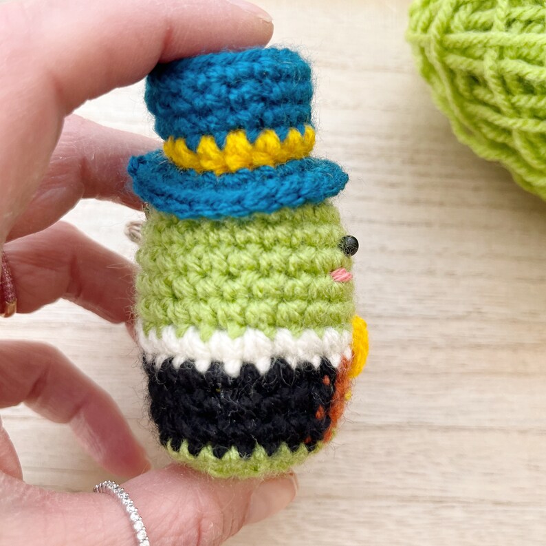 Puede incluir: Una mu&ntilde;eca de ganchillo hecha a mano con un cuerpo verde, pantalones negros y un sombrero de copa azul con ribete amarillo. La mu&ntilde;eca tiene ojos negros peque&ntilde;os y un rubor rosa. La mu&ntilde;eca est&aacute; en una mano.