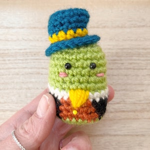 Puede incluir: Una figura de ganchillo hecha a mano de una criatura verde que lleva un sombrero de copa azul con ribete amarillo. La figura tiene ojos negros, mejillas rosadas y una pajarita amarilla. Lleva un traje negro y marr&oacute;n.