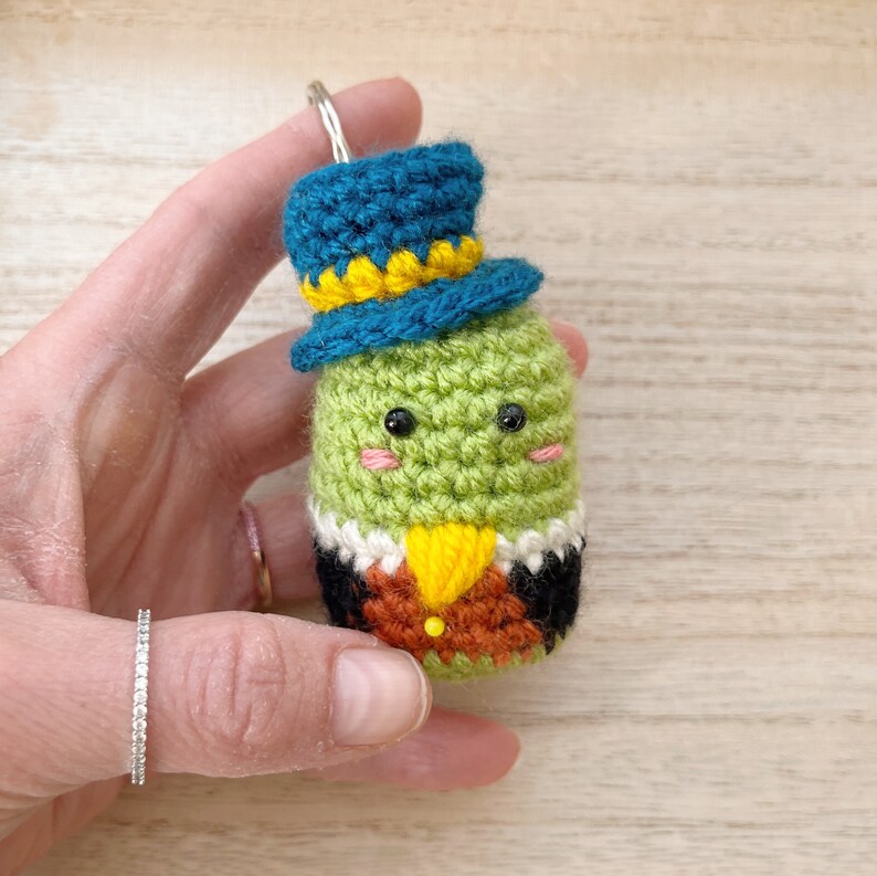 Puede incluir: Un llavero de ganchillo verde hecho a mano con forma de personaje que lleva un sombrero de copa azul con una banda amarilla. El personaje tiene ojos negros, mejillas rosadas y un chaleco amarillo y marr&oacute;n. El llavero est&aacute; sujeto a un anillo plateado.