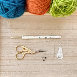 Puede incluir: Un conjunto de suministros de artesan&iacute;a con tres ovillos de lana en turquesa, naranja y verde. Un ganchillo, tijeras doradas, alfileres, una aguja y un marcador de punto est&aacute;n dispuestos sobre una superficie de madera.
