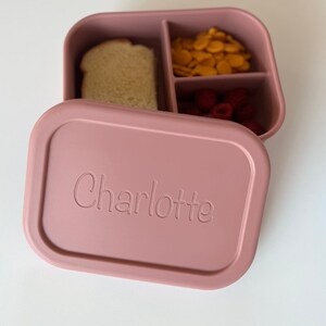 Puede incluir: Una fiambrera de silicona rosa con tres compartimentos. La fiambrera est&aacute; etiquetada con "Charlotte" y contiene un s&aacute;ndwich, galletas y frambuesas.