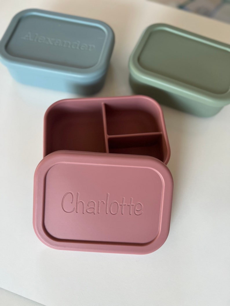 Puede incluir: Dos fiambreras de silicona, una azul con el nombre "Alexander" y otra rosa con el nombre "Charlotte" y un compartimento dividido.