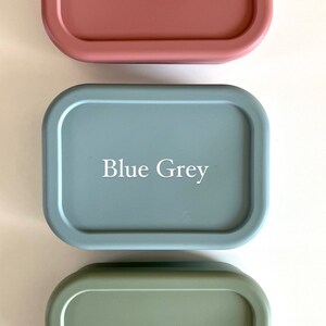 Puede incluir: Tres recipientes rectangulares de silicona con bordes redondeados. Los recipientes son de color rosa blush, gris azulado y verde salvia. Cada recipiente tiene el nombre del color impreso en blanco en la parte delantera.