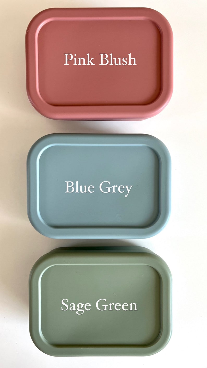 Puede incluir: Tres recipientes rectangulares de silicona con bordes redondeados. Los recipientes son de color rosa blush, gris azulado y verde salvia. Cada recipiente tiene el nombre del color impreso en blanco en la parte delantera.
