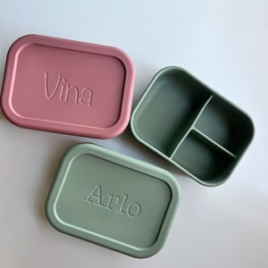 Puede incluir: Dos fiambreras rectangulares de silicona, una de color rosa apagado con el nombre "Vina" grabado en la tapa, y la otra de color verde apagado con el nombre "Arlo" grabado en la tapa. La fiambrera verde tiene un interior dividido.