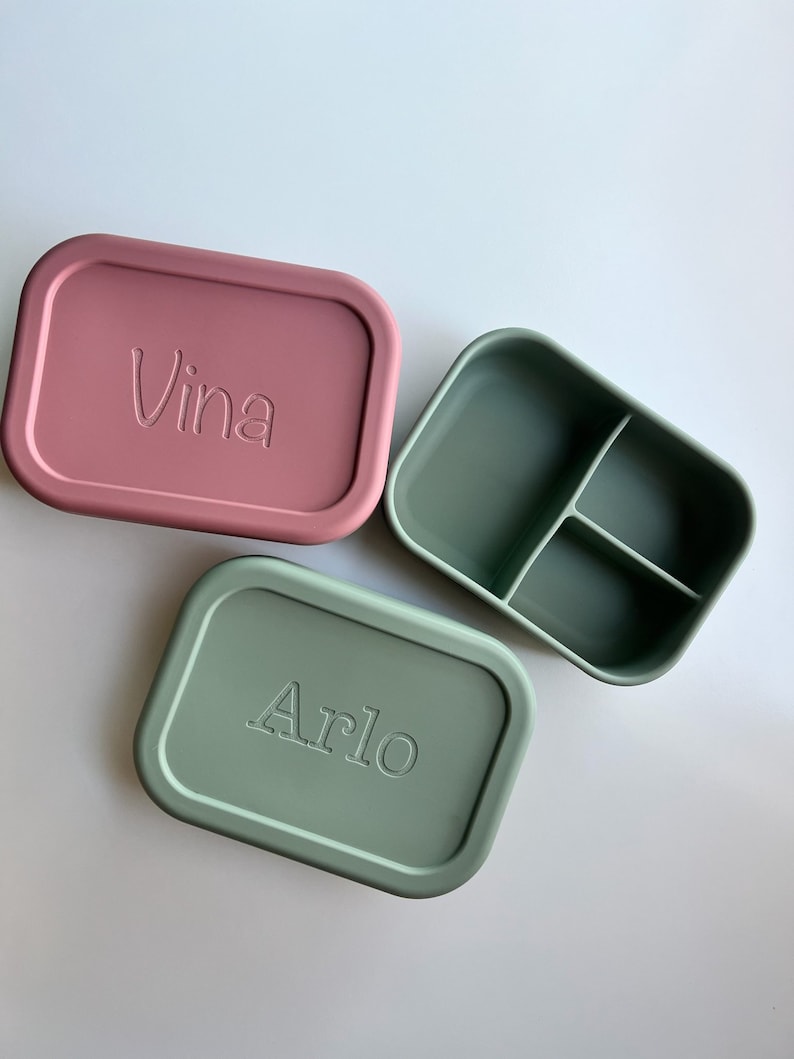 Puede incluir: Dos fiambreras rectangulares de silicona, una de color rosa apagado con el nombre "Vina" grabado en la tapa, y la otra de color verde apagado con el nombre "Arlo" grabado en la tapa. La fiambrera verde tiene un interior dividido.