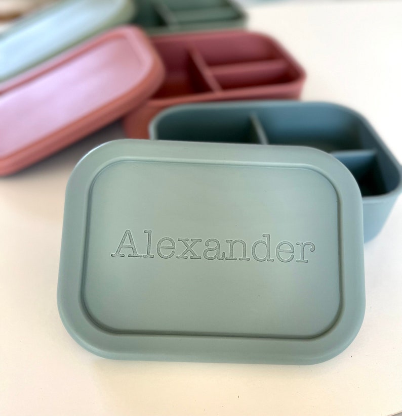 Puede incluir: Una fiambrera de silicona azul verdosa con un interior dividido. La tapa tiene el nombre "Alexander" grabado.