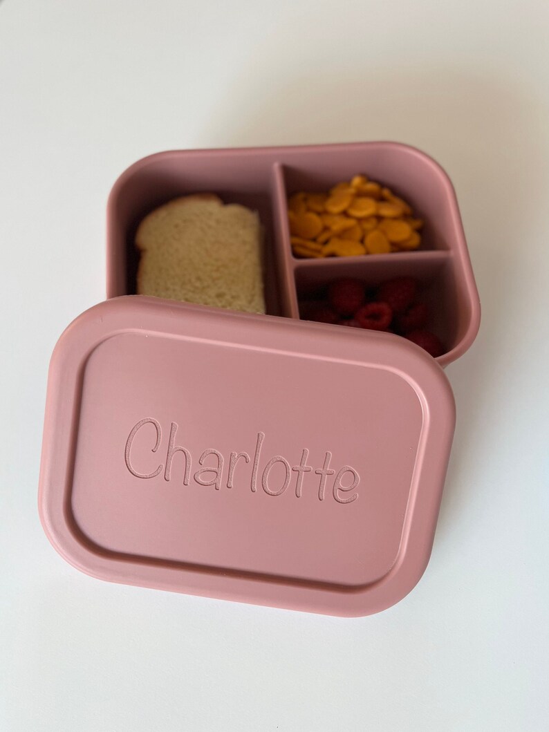 Puede incluir: Una fiambrera de silicona rosa con tres compartimentos. La fiambrera est&aacute; etiquetada con "Charlotte" y contiene un s&aacute;ndwich, galletas y frambuesas.