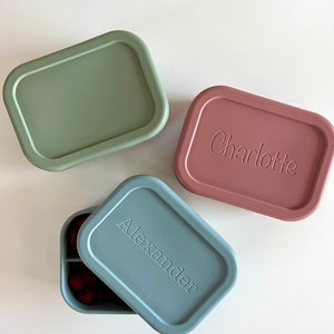 Puede incluir: Tres fiambreras de silicona con nombres personalizados. Las fiambreras son de color verde, rosa y azul. La fiambrera azul est&aacute; abierta y tiene un interior dividido.