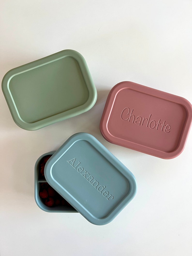 Puede incluir: Tres fiambreras de silicona con nombres personalizados. Las fiambreras son de color verde, rosa y azul. La fiambrera azul est&aacute; abierta y tiene un interior dividido.