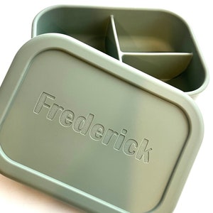 Puede incluir: Una fiambrera rectangular verde con un interior dividido. La tapa tiene el nombre "Frederick" grabado en relieve.