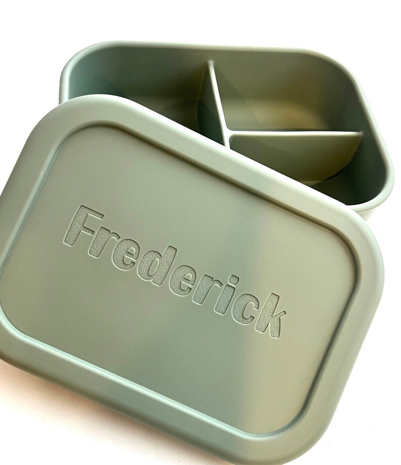 Puede incluir: Una fiambrera rectangular verde con un interior dividido. La tapa tiene el nombre "Frederick" grabado en relieve.