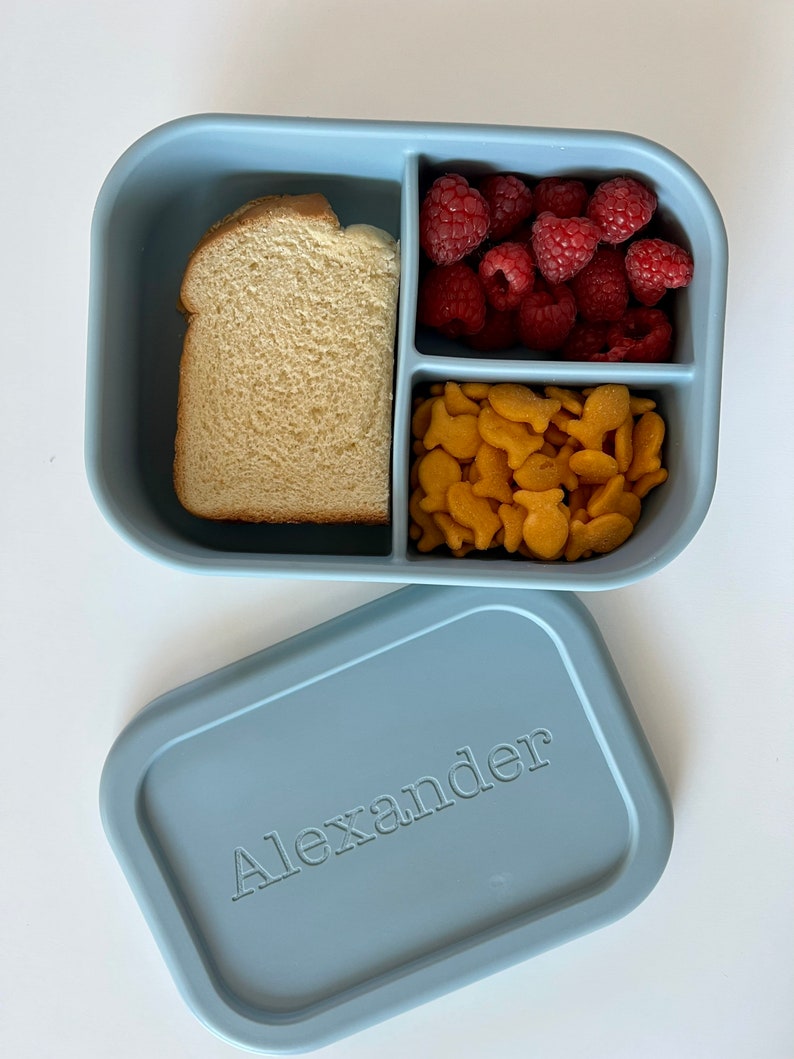 Puede incluir: Una fiambrera de silicona azul con tres compartimentos. Los compartimentos contienen un s&aacute;ndwich, frambuesas y galletas de pescado. La tapa de la fiambrera es azul y tiene el nombre "Alexander" grabado.