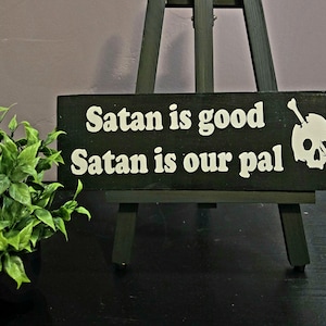 May include: A black wooden sign with white text that reads "Satan is good, Satan is our pal." A white skull with a bone through it is on the right side of the sign. The sign is displayed on a dark green easel.