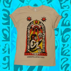 Può includere: Una t-shirt beige con un'illustrazione in nero e giallo di una scimmia che tiene un teschio davanti a un arco rosso, giallo e nero con un grande occhio e raggi di luce. Il testo "SHANEMILLER TATTOOS" è stampato sotto l'illustrazione.