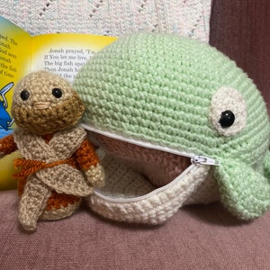 Puede incluir: Una ballena verde de crochet con una abertura con cremallera, que revela un interior de crochet blanco. Una figura marrón de crochet con una túnica beige está sentada dentro de la ballena.