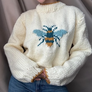 Puede incluir: Jersey de punto color crema con un gran diseño de abeja en la parte delantera. La abeja tiene alas azules y doradas y un cuerpo a rayas. El jersey tiene un cuello alto y puños acanalados con un ribete dorado. Una prenda acogedora y hecha a mano.