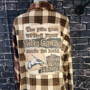 May include: A plaid flannel shirt in cream, brown, and tan. The back features a patch with the text "The pen that writes your life story must be held in your own hand" and an illustration of a hand holding a pen.