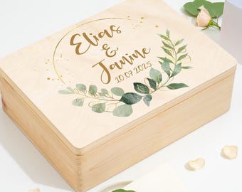 Può includere: Una scatola di legno color chiaro con coperchio incernierato. Il coperchio è decorato con un design circolare color oro, i nomi "Elias & Janine" e la data "10.07.2025". Accenti floreali verdi e bianchi circondano il testo. La scatola è rettangolare.