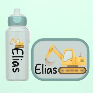 Könnte beinhalten: Eine hellgraue Wasserflasche und eine passende Brotdose. Beide Artikel zeigen ein gelbes Bagger-Design und den Namen "Elias" in Schwarz. Die Flasche hat einen teal-farbenen Deckel und eine weiße Trageschlaufe. Die Brotdose ist rechteckig.