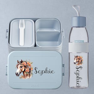 Könnte beinhalten: Ein hellblaues Lunchbox- und Wasserflaschen-Set. Die Lunchbox zeigt eine Pferdeillustration mit floralen Akzenten und dem Namen "Sophie". Die Wasserflasche hat ein passendes Design. Das Set enthält eine weiße Gabel.