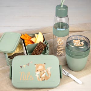 Könnte beinhalten: Ein salbeigrünes Lunch-Set bestehend aus einer rechteckigen Bento-Box, einer transparenten Trinkflasche mit grünem Deckel und einem runden Snackbehälter. Alle Teile sind mit einem Fuchsmotiv und dem Namen Nils bedruckt. Eine kleine Gabel liegt bei.
