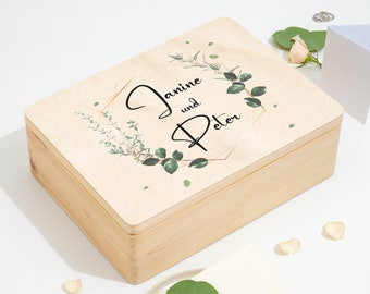 Può includere: Una scatola di legno con una finitura in legno chiaro e un coperchio con un motivo floreale con foglie verdi e accenti dorati. Il coperchio è personalizzato con i nomi "Janine e Peter" in lettere nere.