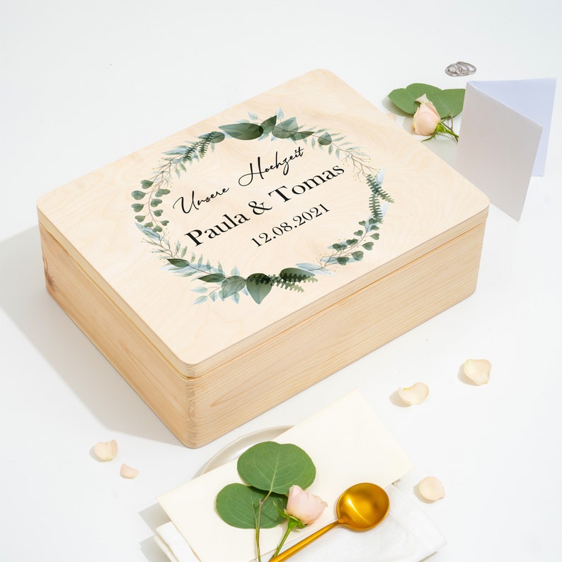 Puede incluir: Una caja de madera con una tapa que presenta una corona de acuarela verde con el texto "Unsere Hochzeit Paula & Tomas 12.08.2021".