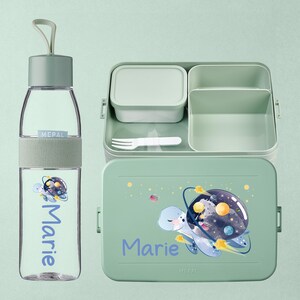Pode incluir: Um conjunto de almoço verde claro com uma garrafa de água a condizer. A lancheira apresenta um design de tartaruga com planetas e o nome "Marie". O conjunto é de plástico.