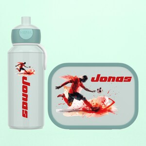 Könnte beinhalten: Ein hellblaues Trinkflaschen- und Brotdosen-Set. Die Trinkflasche und die Brotdose zeigen eine Grafik eines Fußballspielers mit dem Namen "Jonas" in Rot. Die Trinkflasche hat einen weißen Riemen und einen hellblauen Deckel.
