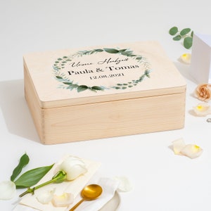 Puede incluir: Una caja de madera con una tapa que presenta un dise&ntilde;o de corona de hojas verdes y el texto "Unsere Hochzeit Paula & Tomas 12.08.2021".