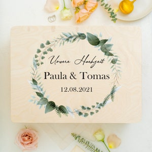 Puede incluir: Un letrero de madera con un dise&ntilde;o de corona floral verde y blanca. El letrero dice "Unsere Hochzeit" (Nuestra boda) Paula & Tomas 12.08.2021.