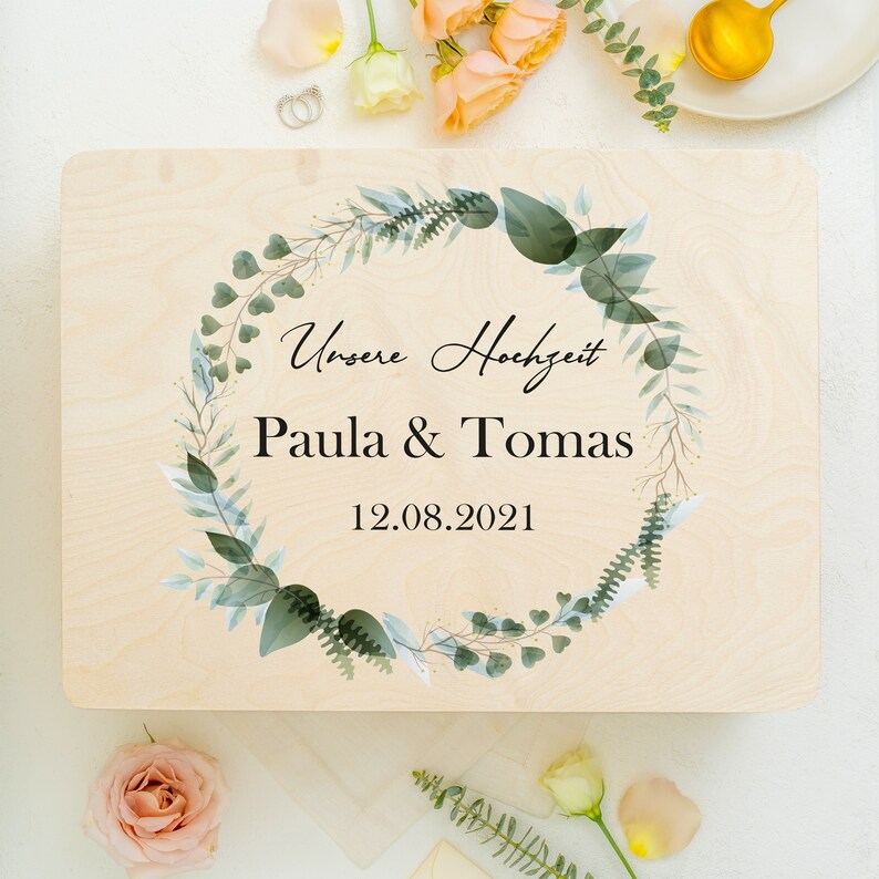 Puede incluir: Un letrero de madera con un dise&ntilde;o de corona floral verde y blanca. El letrero dice "Unsere Hochzeit" (Nuestra boda) Paula & Tomas 12.08.2021.
