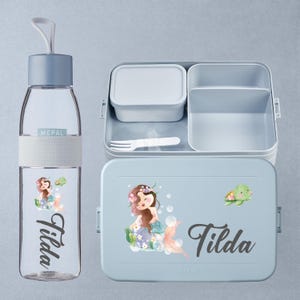 Pode incluir: Conjunto Mepal azul claro com garrafa de água e lancheira. A garrafa tem uma tampa azul clara e uma faixa branca. A lancheira tem um desenho de sereia e tartaruga, com o nome "Tilda" em itálico. Inclui um recipiente pequeno e um garfo.