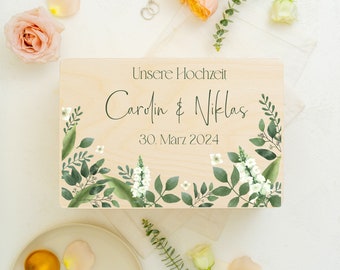 Può includere: Una scatola di legno con un design floreale e il testo "Unsere Hochzeit Carolin & Niklas 30. März 2024" sul coperchio.