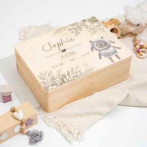 Può includere: Una scatola portaoggetti in legno con una finitura in legno chiaro. Il coperchio presenta un design ad acquerello con foglie verdi e un adorabile agnello bianco. La scatola è personalizzata con il nome "Sophie", la data "29.10.2024", l'ora "18:30 Uhr", il peso "3510 g" e la lunghezza "44 cm".