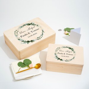 Puede incluir: Dos cajas de madera con tapas, decoradas con una corona de hojas verdes y el texto "Unsere Hochzeit Paula & Tomas 12.08.2021".