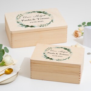 Puede incluir: Dos cajas de madera con tapas, ambas decoradas con una corona de hojas verdes y el texto "Unsere Hochzeit Paula & Tomas 12.08.2021".