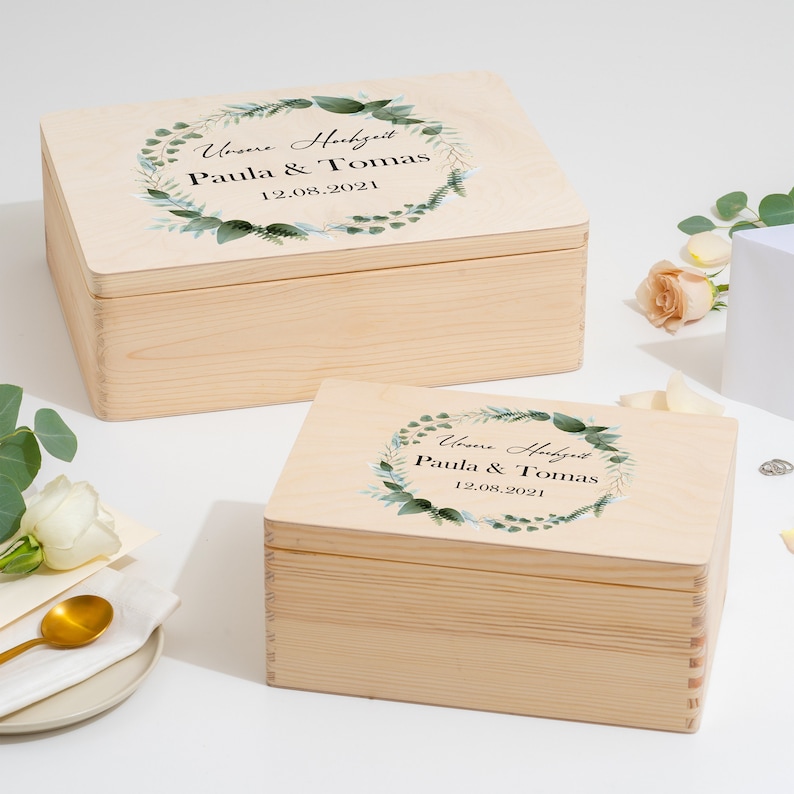 Puede incluir: Dos cajas de madera con tapas, ambas decoradas con una corona de hojas verdes y el texto "Unsere Hochzeit Paula & Tomas 12.08.2021".