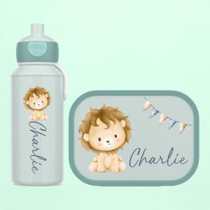 Könnte beinhalten: Ein hellblaues Wasserflaschen- und Lunchbox-Set. Beide Artikel zeigen eine Cartoon-Löwen-Illustration und den Namen "Charlie" in Schreibschrift. Die Lunchbox hat ein Banner mit kleinen Flaggen. Die Wasserflasche hat einen hellblauen Deckel.