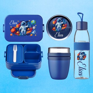 Könnte beinhalten: Ein blaues Lunch-Set mit Astronauten-Design und dem Namen "Elias". Das Set enthält eine Lunchbox, einen runden Behälter, eine Wasserflasche und einen Essensbehälter mit einer Gabel. Das Astronauten-Design zeigt Planeten und eine Rakete.