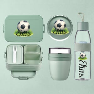 Könnte beinhalten: Ein Set grüner Lunch-Accessoires mit Fußball-Design. Enthält eine Brotdose, einen runden Behälter, einen Lebensmittelbehälter und eine Wasserflasche. Der Name "Elias" ist auf der Brotdose und der Wasserflasche aufgedruckt.