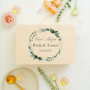 Puede incluir: Una caja de madera con un dise&ntilde;o de corona floral verde y blanca. La caja tiene el texto "Unsere Hochzeit" y "Paula & Tomas" con la fecha "12.08.2021".