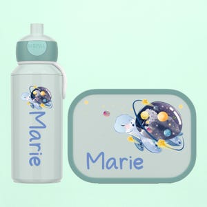Könnte beinhalten: Ein hellgrünes Wasserflaschen- und Lunchbox-Set. Die Flasche und die Lunchbox zeigen eine Zeichentrickschildkröte mit einem Galaxie-Schalen-Design. Der Name "Marie" ist in Hellblau auf beiden Artikeln aufgedruckt.