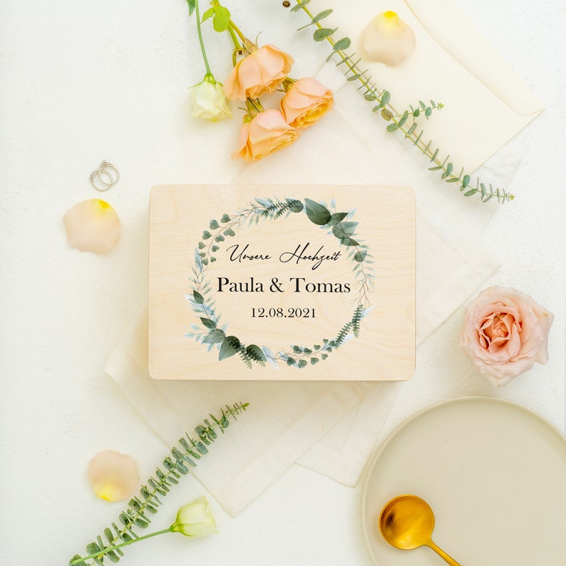 Puede incluir: Una caja de madera con un dise&ntilde;o de corona floral y el texto "Unsere Hochzeit Paula & Tomas 12.08.2021" en la tapa.