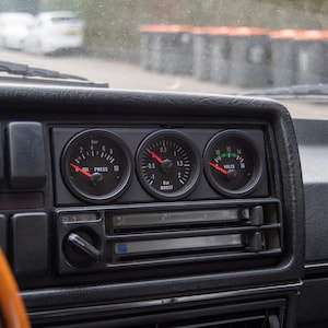 Può includere: Primo piano del cruscotto di un'auto con tre indicatori neri. Gli indicatori misurano la pressione dell'olio in bar, la pressione di sovralimentazione in bar e la tensione in volt. Gli indicatori hanno lancette bianche e marcature bianche.