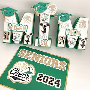 May include: Green, white and gold letter blocks spelling "MITM" with cheerleading-themed decorations. Each letter has a gold glitter graduation cap, a black and white cheerleader silhouette, and a gold and white "Senior Cheer 2024" banner. The letters are arranged on a green and gold sign that reads "SENIORS Cheer 2024".