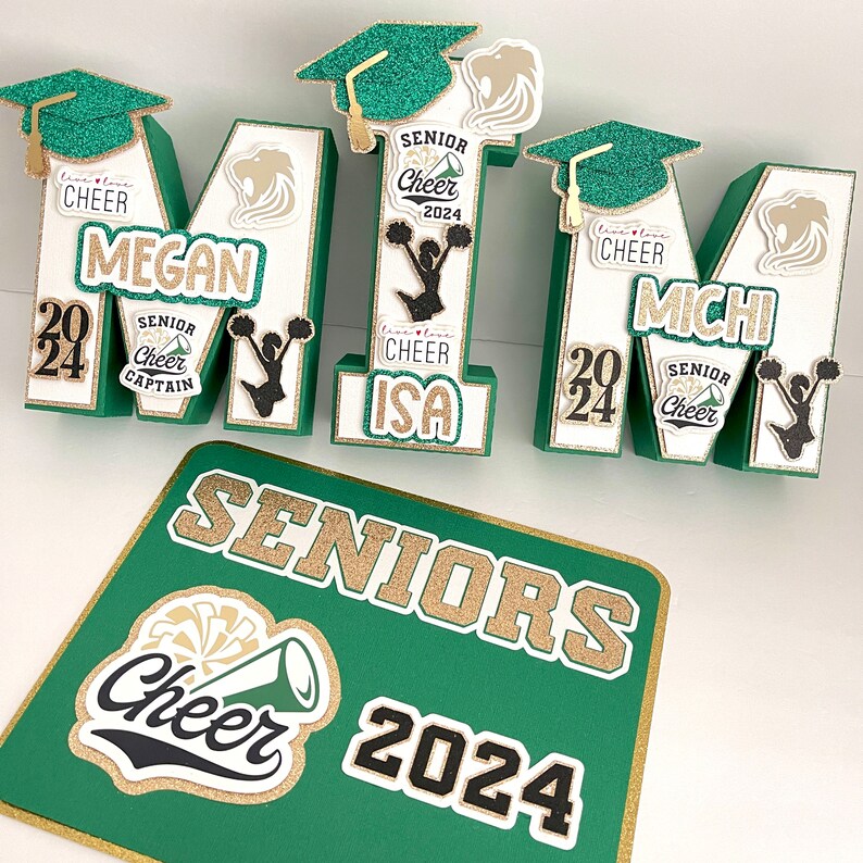 May include: Green, white and gold letter blocks spelling "MITM" with cheerleading-themed decorations. Each letter has a gold glitter graduation cap, a black and white cheerleader silhouette, and a gold and white "Senior Cheer 2024" banner. The letters are arranged on a green and gold sign that reads "SENIORS Cheer 2024".